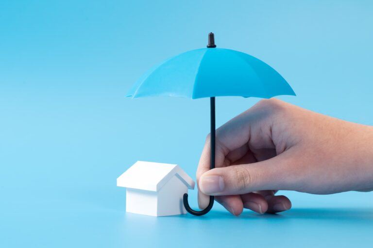 A hand holds a small blue umbrella over a tiny white model of a house, representing mortgage protection.