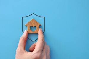 A hand places a small wooden house inside the outline of a shield, symbolising mortgage protection.