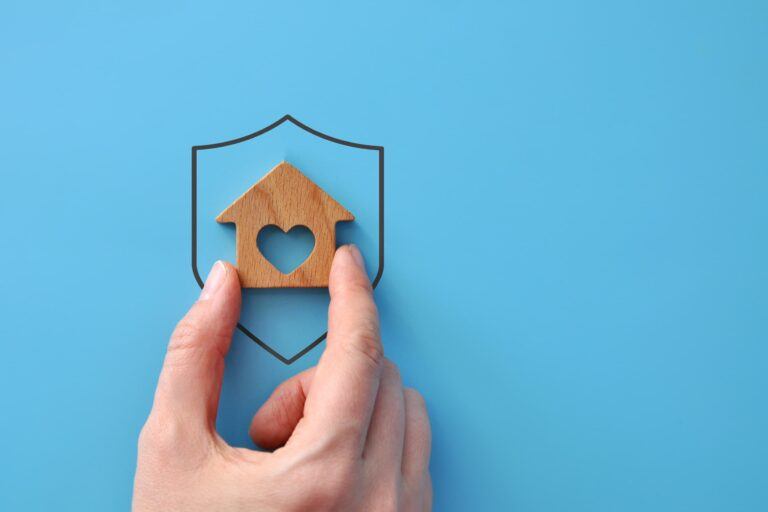 A hand places a small wooden house inside the outline of a shield, symbolising mortgage protection.