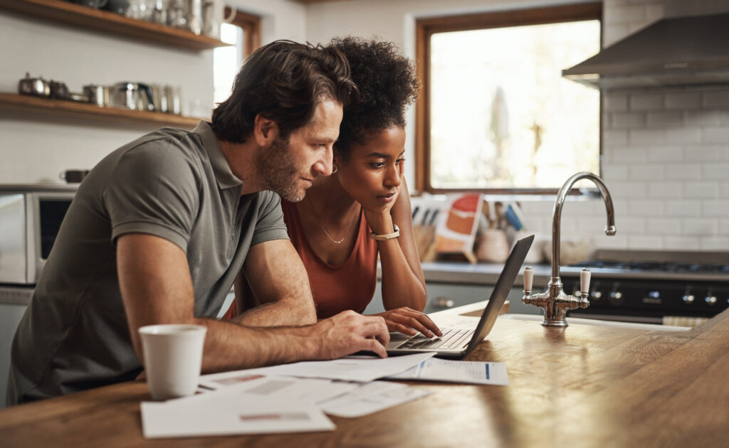 A couple looking at a laptop and paper documents financial planning in a kitchen.